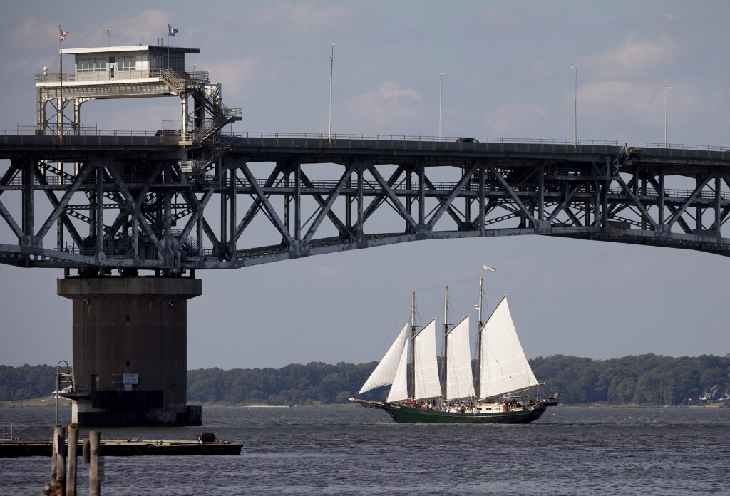 A three-masted tall ship with white sails passes beneath the George P. Coleman Memorial Bridge spanning the York River in Virginia on a partly cloudy day.